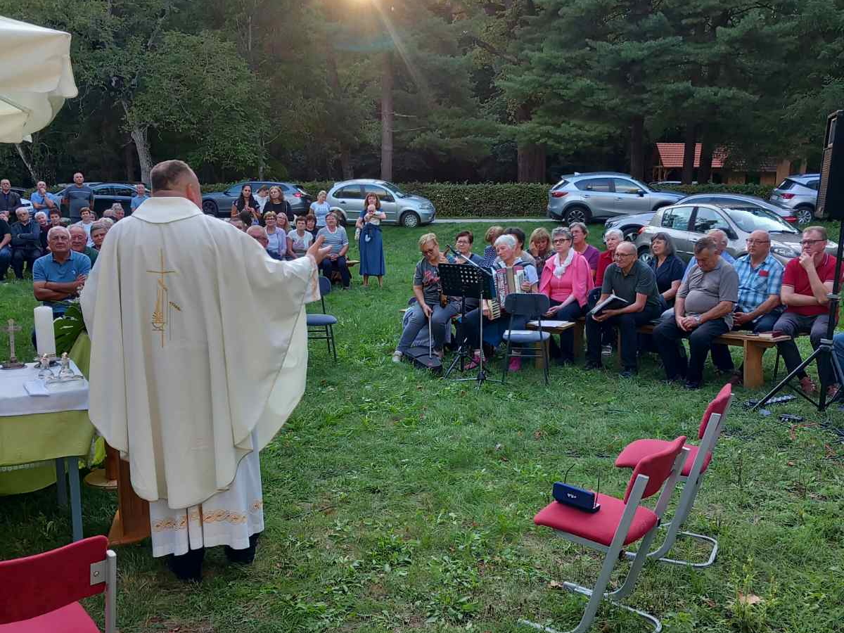 Misa na otvorenom u Križančiji - svećenik u bijeloj kazuli pred vjernicima koji sjede na klupama u šumi pri zalasku sunca.